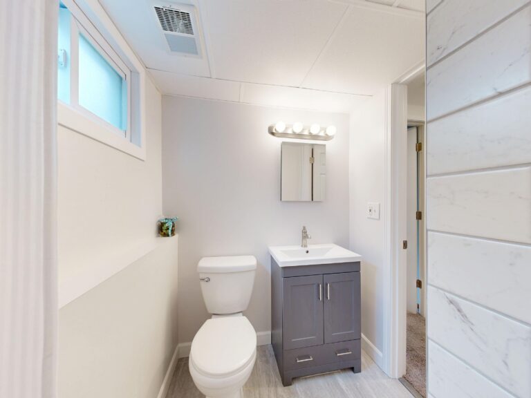 Compact finished basement bathroom with a grey vanity, toilet and a small high window near the ceiling
