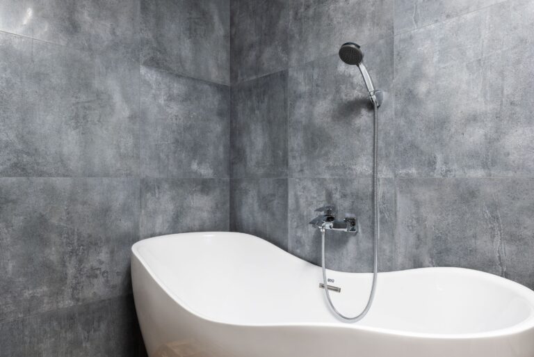 Freestanding bathtub with a hand shower against a large-format grey tiled wall